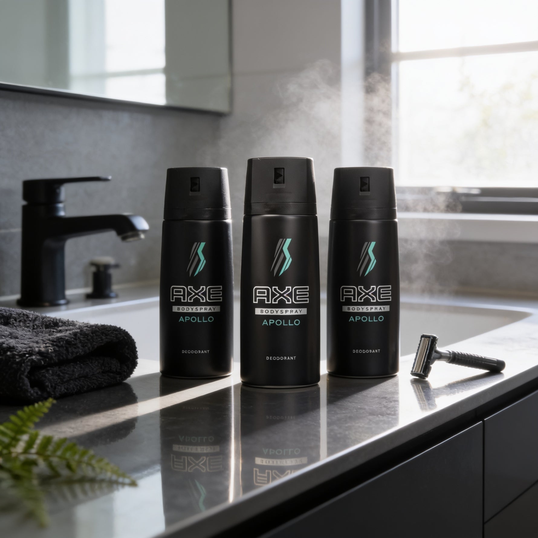 Three black deodorant cans with theAXE brand name and the word 'APOLLO' printed on them, placed on a bathroom counter with a razor and a black towel.