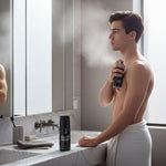 A young man stands in a bathroom, holding a can of deodorant and spraying it on his back.